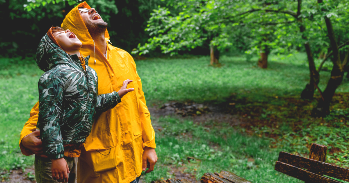 A father and son stand outside in raincoats catching raindrops in their mouths.