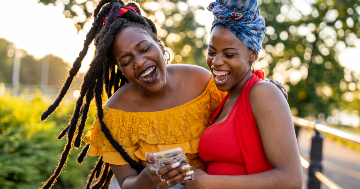 Two friends stand outside looking at a cell phone and laughing.