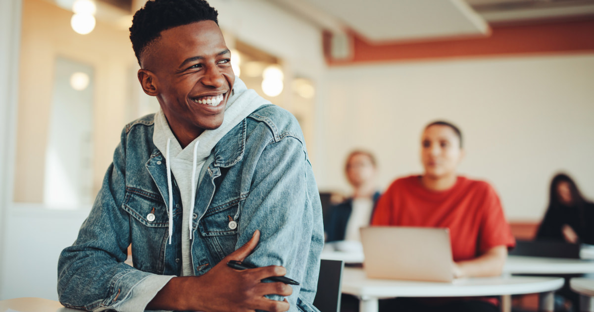 A college-age man sits at a desk and smiles