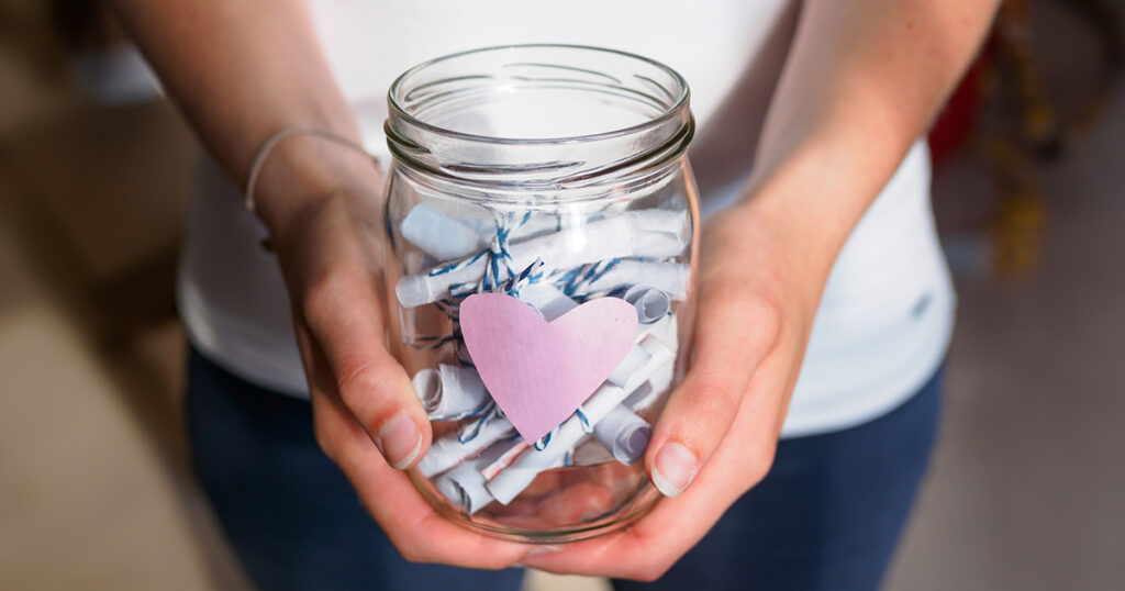 Two hands hold a mason jar filled with rolled up notes tied with string. There is a pink paper heart on the front of the jar.