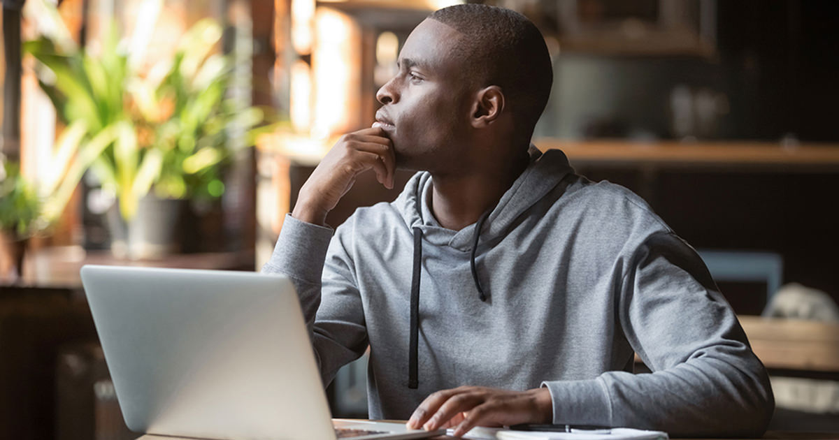 A guy looking out a window sits in front of an open laptop computer.