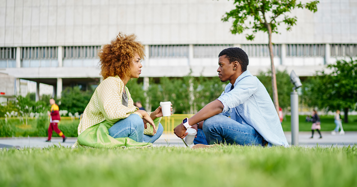 A man and a woman sit in the grass outside a building talking.