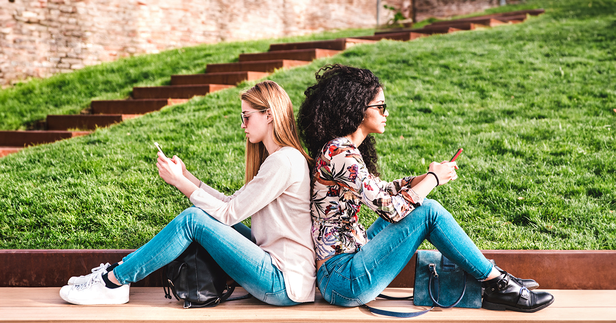 Two women using their cell phones sit with their backs touching.