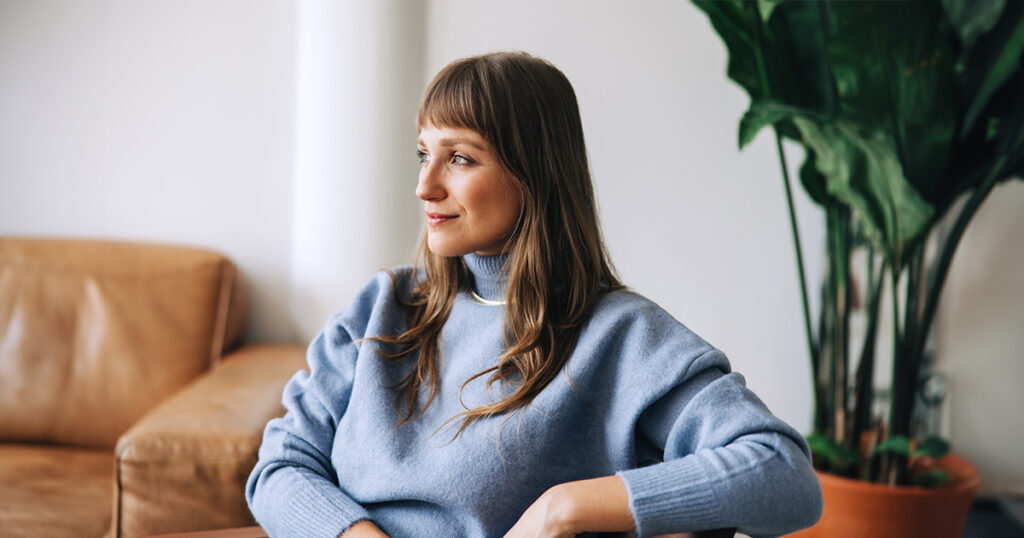 A woman in a periwinkle turtleneck sitting in a lounge area looks off into the distance.