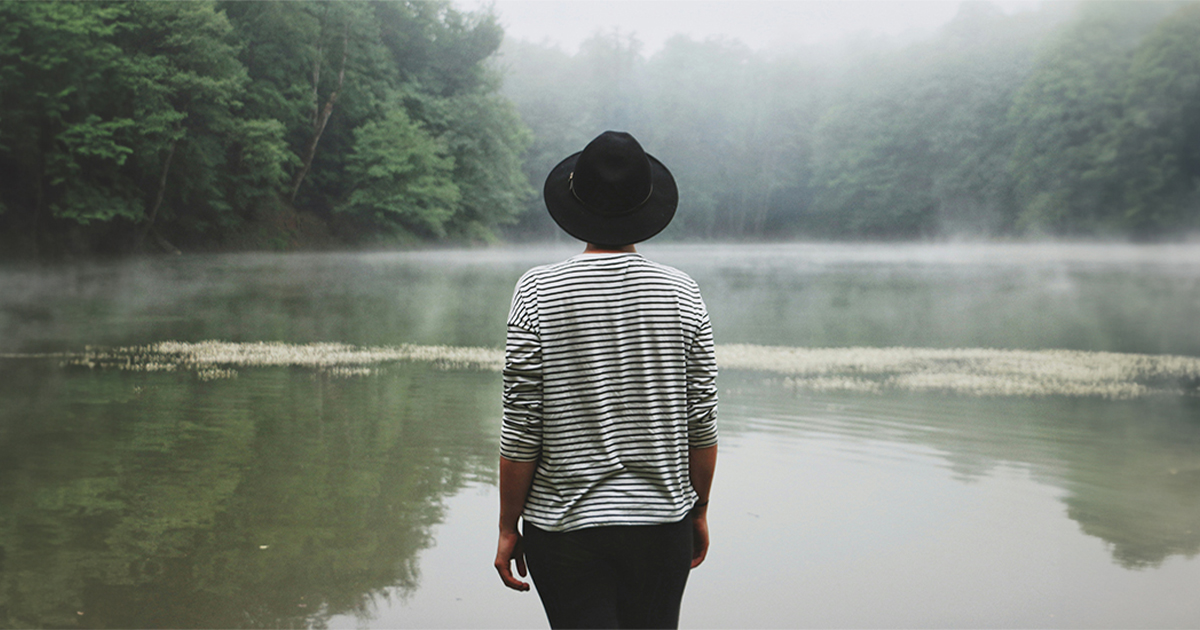 A person in a black hat looks at the mist rising from pond.