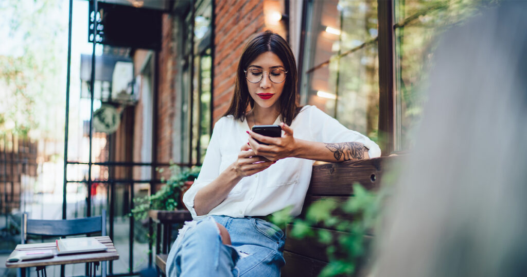 A woman sitting outside on a bench looks at her phone.